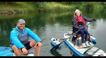 A man on an adapted seat paddleboard and a lady with a wheelchair adapted paddleboard with extra floats for stability on a lake