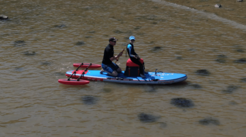 Boy and a man on a paddleboard in the sea, the boy is sitting on an adaptable chair whilst the man is paddling behind him. Floats have been added to the paddleboard for stability.