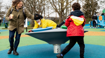 A lady and a boy spinning another boy on an accessible metal spinning top in the park