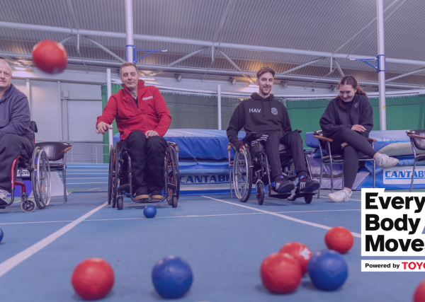 A group of 3 white males playing Boccia sat in manual wheelchairs with a white female sat next to them smiling as one of the men throws a red Boccia ball towards the camera, which is slightly blurred and in mid air as it's about to land in the middle of a set of red and blue Boccia balls.