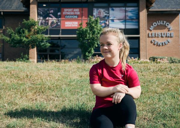 Paralympics swimmer Maisie Summers-Newton sitting on a grassy bank outside a leisure centre.