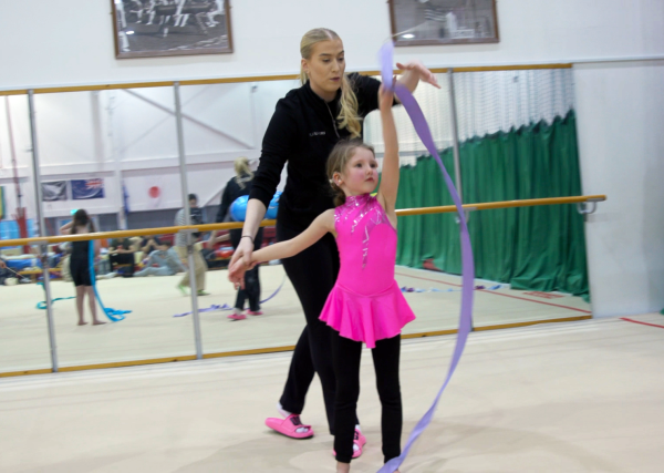 A young girl dressed in pink and black gymnastics outfit, taking part in rhythmic gymnastics. She is holding the ribbon above her head and is supported from the back by her coach dressed in black. They are participating inside a gymnastics hall, with padded floor and mirrored walls.