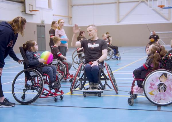 Rhyl Raptors COTM main story cover - 3 wheelchair users, 1 coach in the middle and 2 young girls each side, engaged in a wheelchair basketball training session on an indoor court.