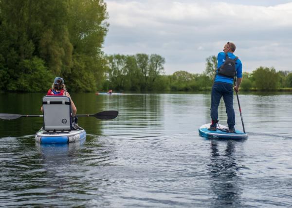 The back of a man and a woman paddleboarding on a lake. The woman is in an adapted seat whilst the man is accompanying her stood up.