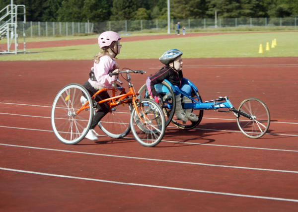 two children in wheelchair athletics chairs on a red athletics track with a green field in the background.