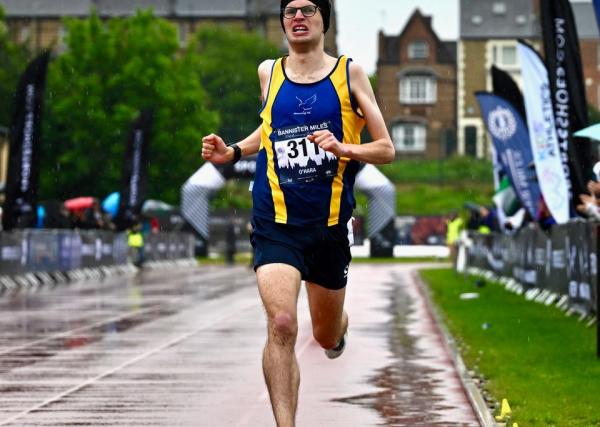 Kieran O'Hara running through a wet backdrop on a running track wearing blue running vest.