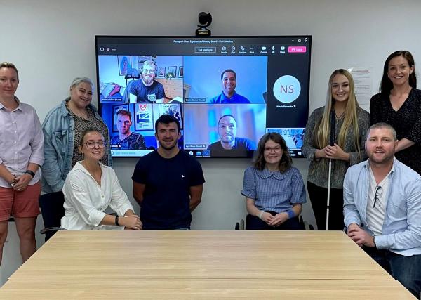 The Lived experience advisory board are pictured in a hybrid meeting with attendees in person and online pictured in a group around a large screen.