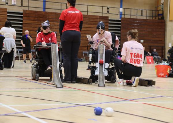 Group of boccia players mid game, sat in chairs facing the camera and using ramp launch aids