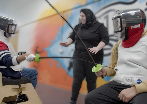 2 seated fencers mid bout, with swords touching and dressed in full protective gear.