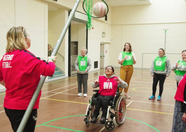 Inclusive netball session on an indoor court. Wheelchair participant taking a shot at a net being held by a coach in the foreground