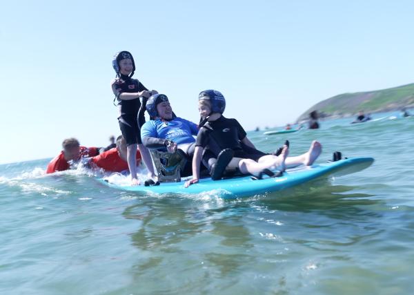 A group of children and a man using adaptive equipment enjoying an inclusive surfing session on a sunny day, supported by volunteers in the sea, with a scenic coastline in the background.