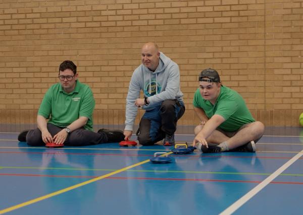 3 partcipants sat on the floor of an indoor sports hall, playing floor curling.