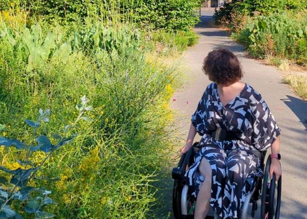 Meg, sitting in her wheelchair surrounded by lush green plants and foliage, in an inner city park 