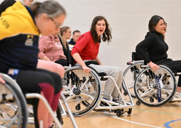 A young woman in a red top and grey tracksuit bottoms takes part in a wheelchair rugby league session on an indoor court. She looks animated and engaged as she pushes her chair forward, surrounded by other participants also using sports wheelchairs. A group of people watch from the sidelines in the background.