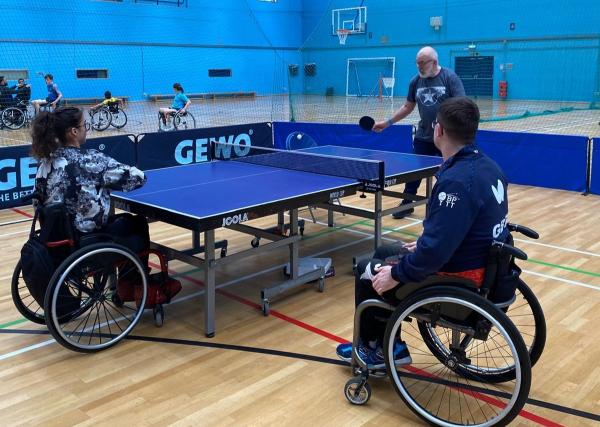Wheelchair users play table tennis in an indoor sports hall, supported by a coach in an accessible and inclusive setting.