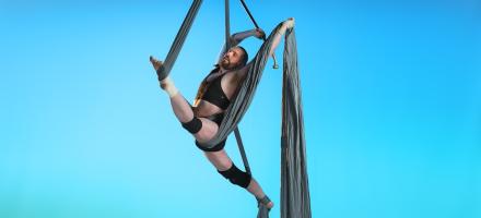 Joel, a non binary person with a masc body is on metallic grey silks with a light blue background. They are wearing very little: black briefs and a black sports bra. They are in a stunning over-split, wrapped carefully within the silks, showing off a variety of supports: ankle, wrist, mechanical knee brace & back brace, holding their brass knobbed walking stick triumphantly above their head smiling softly.